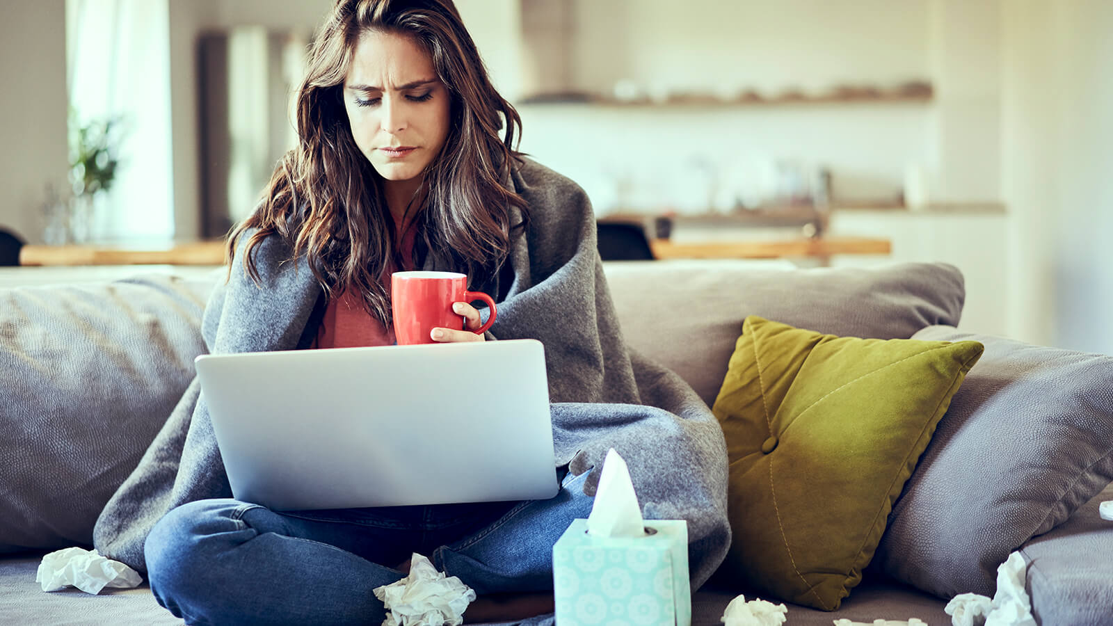 A woman with the flu sits on the couch and works on her laptop