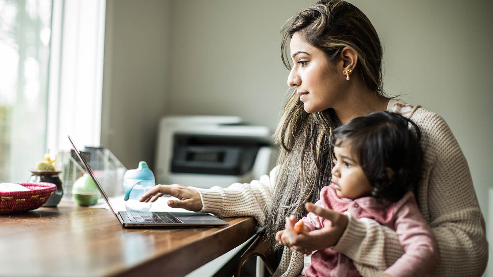 A woman works on her laptop while holding her child